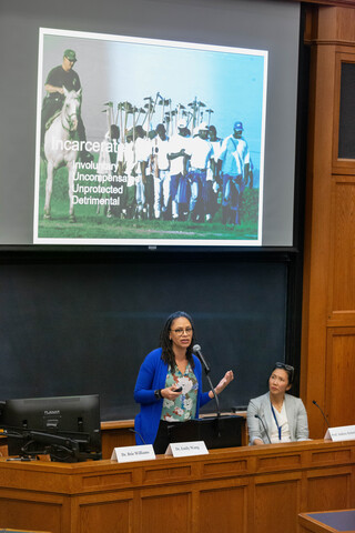 Woman standing at a podium next to a seated woman, both in front of a chalkboard. Above them is a projection screen with a photo of group of men working in a field and the words "Incarcerated: involuntary, uncompensated, unprotected, detrimental."