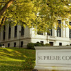 A stone marker with the engraved words “SUPREME COURT” sits in front of a white stone courthouse, surrounded by a lawn and trees 