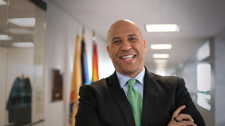 Cory Booker smiles with his arms folded and an office in soft focus behind him.