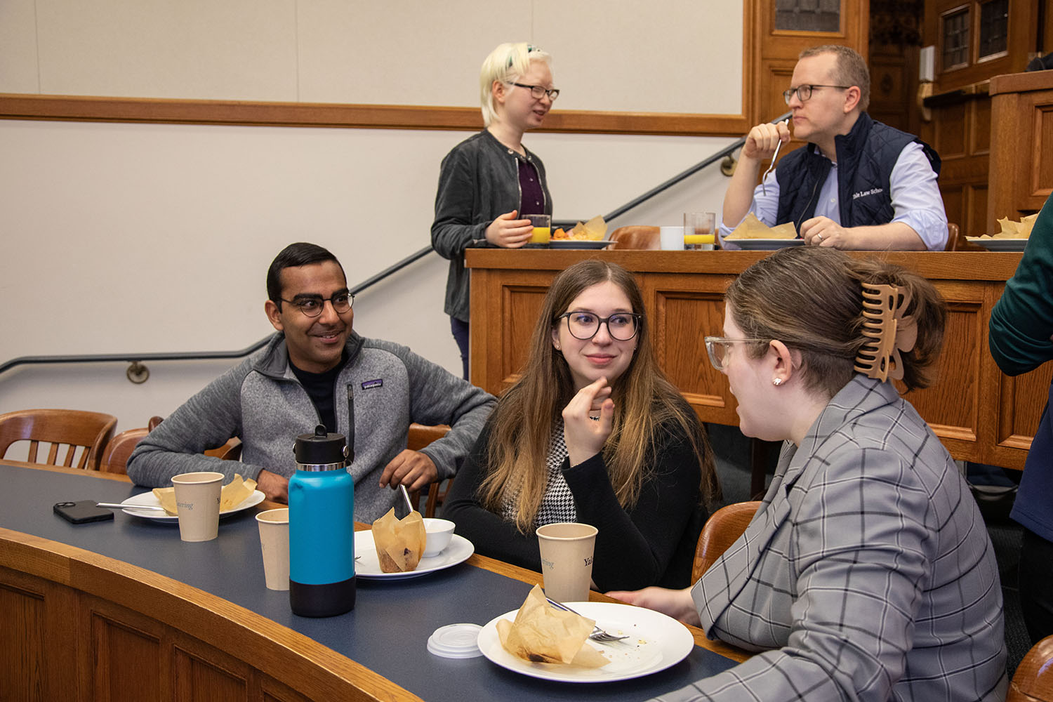 Students sitting at a table with coffee cups and plates of food as one talks.