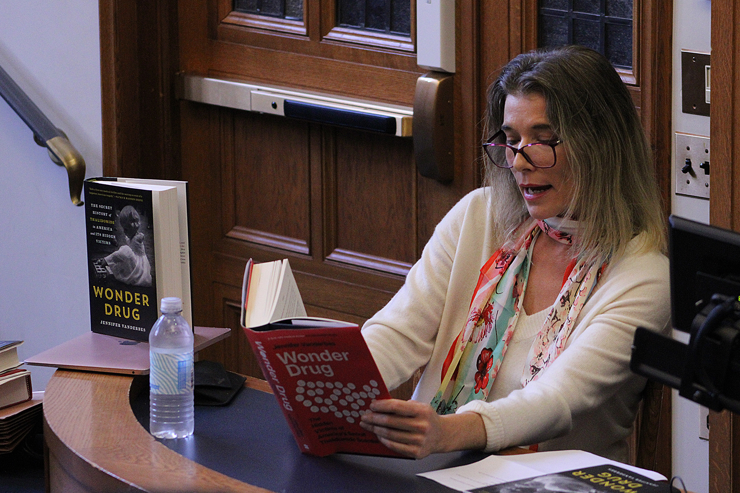 A woman wearing reading glasses, sitting at a classroom desk, reads from a book with the title Wonder Drug. Another edition of a book with the same title is propped on the desk.