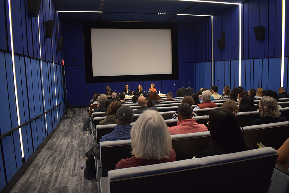 A group of panelists seated at a table before a blank screen, rows of audience members in theater seating