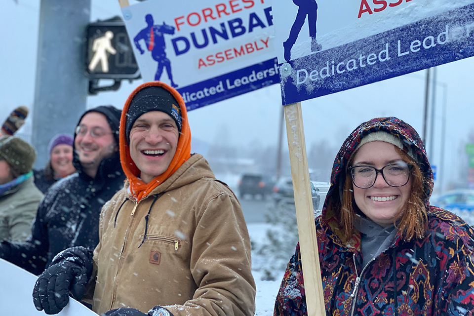 Forrest Dunbar stands outside, while it's snowing, with people holding campaign signs. 