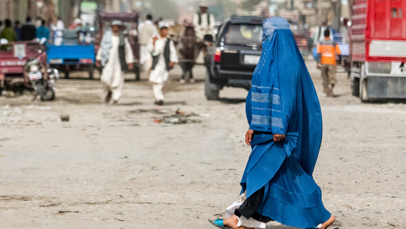 Two women wearing burqa cross a busy unpaved street in Kabul, Afghanistan, with vehicles and other pedestrians visible in the background
