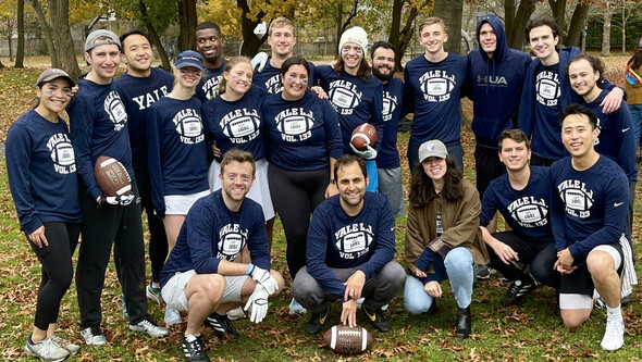 A group of students wearing dark blue Yale t-shirts pose in a park with a football on the ground in front of them