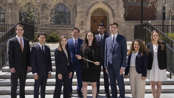 A group of 8 students in suits stand outside the Law School. The female student in the center holds a baton.