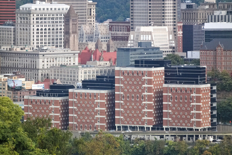 Four squat, blocky brick buildings of varying heights are along a river surrounded by office and other tall buildings in a downtown area
