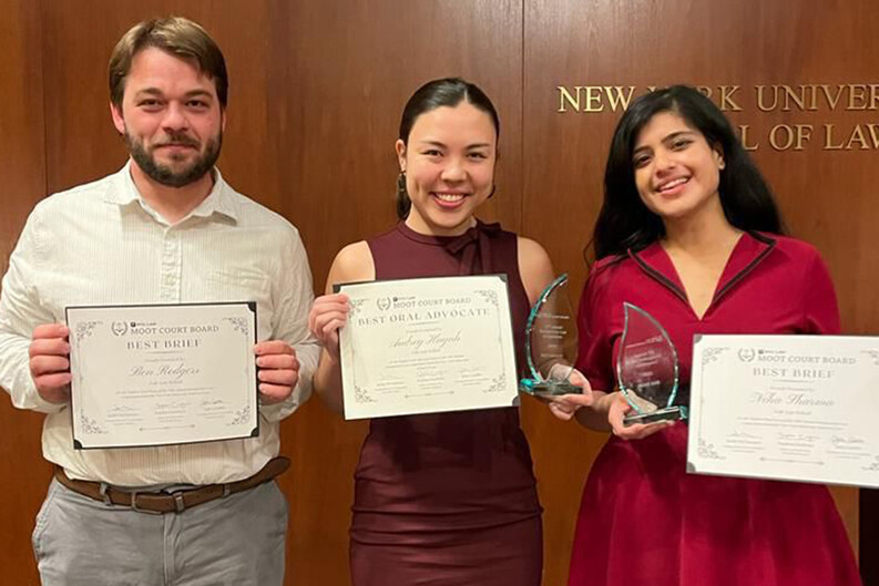 Three students hold up certificates and glass trophies, standing in front of a wood-paneled wall with the words "New York University School of Law"