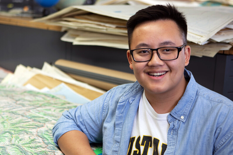A student poses for a photo seated in front of a table piled with maps