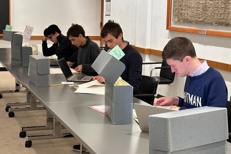 Students, seated at long table with a row of archival file boxes, look at papers.