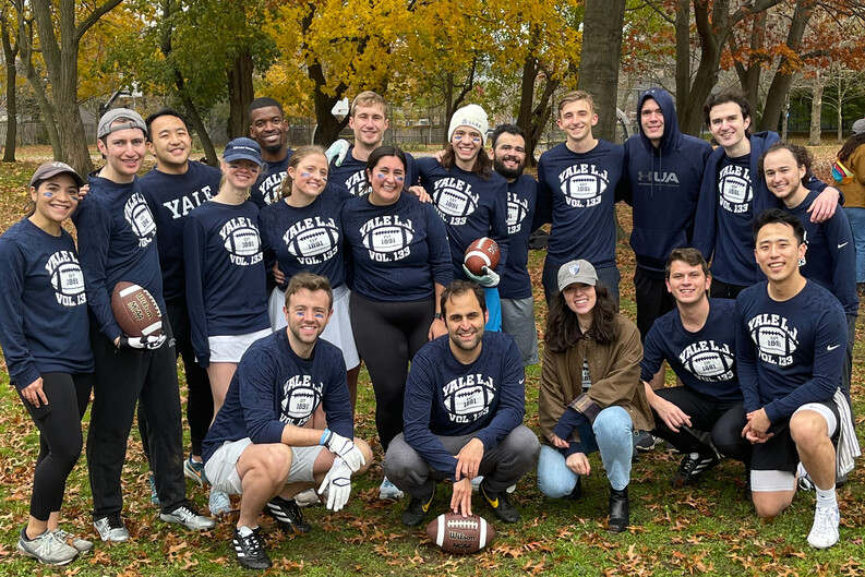 Students wearing team jerseys, standing in a group with a football, on grass covered with fall leaves and colorful foliage in the background