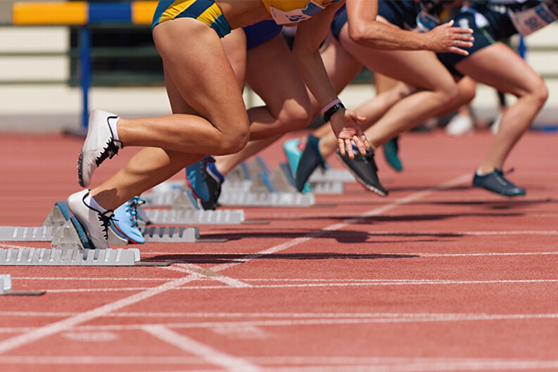 Female runners legs as they leave the starting blocks of a race, running on a reddish track with white stripes 