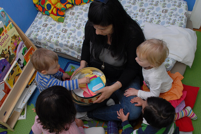 A group of young children and a childcare educator seated in a circle on the floor, looking at colorful papers