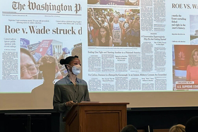 Khiara Bridges at podium before a projection screen with headlines about Dobbs v. Jackson Women’s Health Organization and other abortion-related court cases.