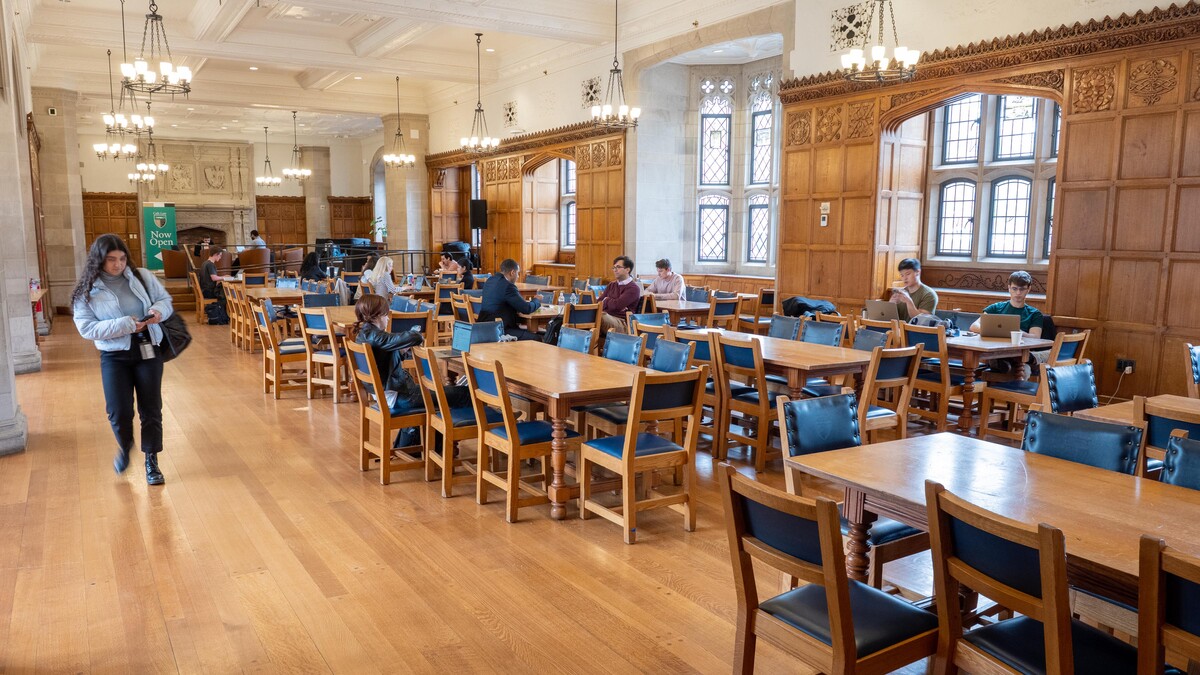 A wide view of Ruttenberg Dining Hall showing students seated at wooden tables eating and studying, with wood paneled walls and large windows 