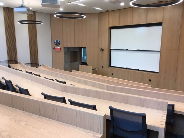 A large tiered lecture hall with blonde wood desks, modern chandeliers, and projection screens.