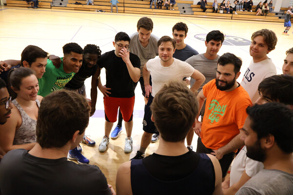 Group of students in gym clothes standing in a circle on the edge of a basketball court, leaning in to listen. 