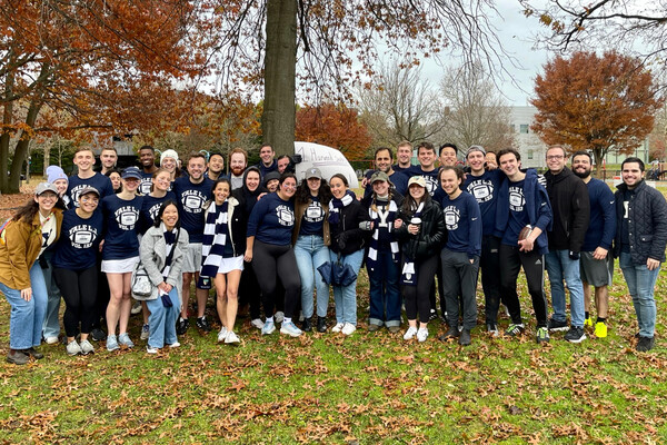 Large group of students wearing Yale jerseys and scarves, stand outside in a park surrounded by fall foliage