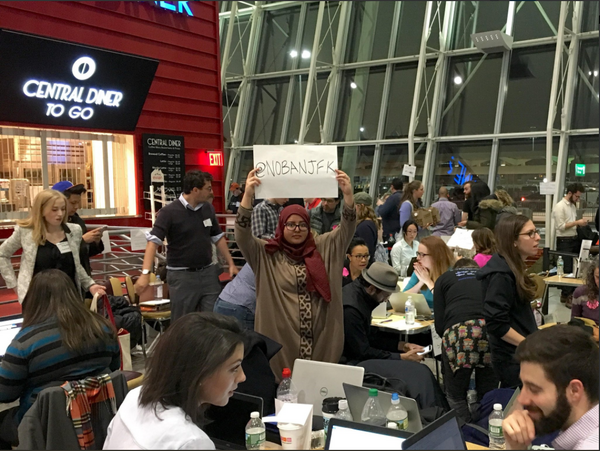 A woman standing in a crowded airport cafe holding a sign reading No Ban JFK