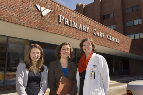 Three women stand in front of a brick building with a sign reading Primary Care Center. One woman wears a white doctor's coat.
