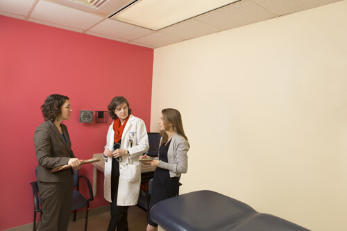 Three women stand in a patient exam room, one wears a white doctor's coat