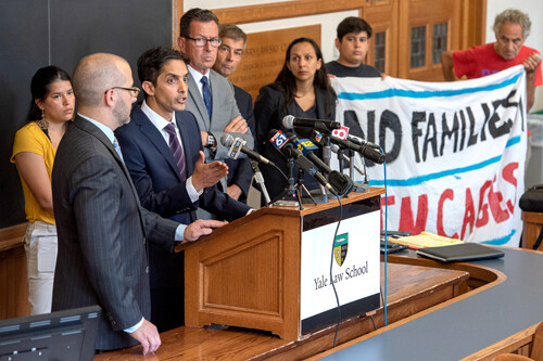 Muneer Ahmad speaking at a podium holding TV station microphones. Next to him is Governor Dannell Malloy and others holding a banner reading No Families in Cages.