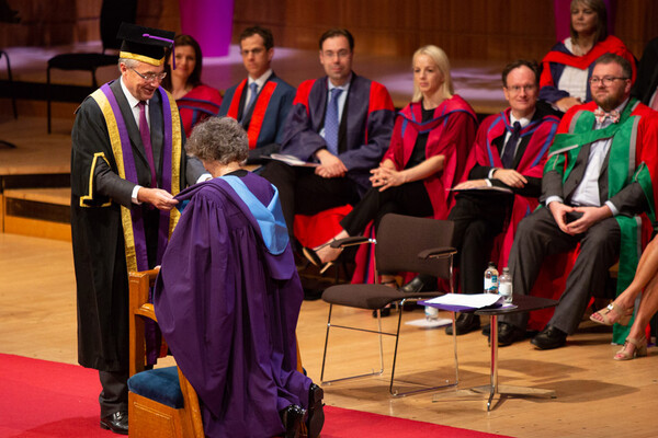 A woman in a purple academic gown kneels before a man presenting her academic hood while onlookers are seated in the rear.
