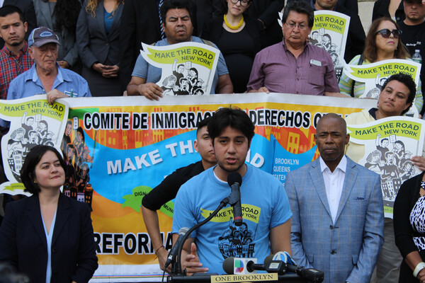 A man speaking at a microphone with people holding a banner behind him reading Make the Road New York