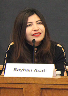 Rayhan Asat speaks into a microphone with a name card on the table in front of her