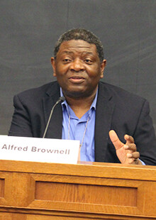 Alfred Brownwell speaks into a microphone with a name card in front of him on a table