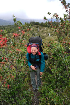 Hannah Duncan with a backpack, crouching on a brushy trail surrounded by flowering bushes, mountains in the background