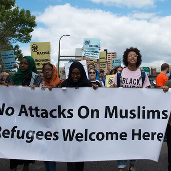 Six people, at the front of a protest, hold a banner that reads "No Attacks on Muslims - Refugees Welcome Here."