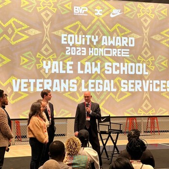Mike Wishnie speaks into a microphone with a screen behind him reading Equity Award 2023 Honoree Yale Law School Veterans Legal Services Clinic