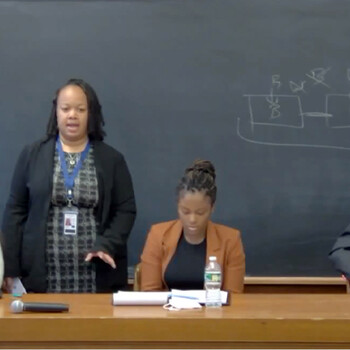 Three women and a man sit at a desk with a blackboard behind them. One woman is standing and speaking.