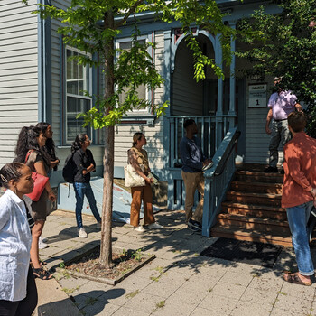 A group of people stand on a sidewalk in front of a house listening to a man talking on the front porch