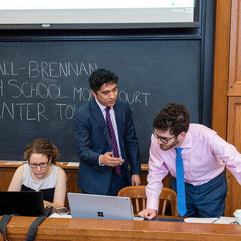 Three students seated at the front of a classroom with a blackboard behind them reading Marshall-Brennan High School Moot Court