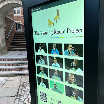 A touch-screen display with "The Visiting Room Project" on the screen with the stone facade of Sterling Law Building in the background