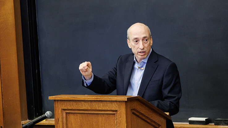 Gary Gensler speaks at a podium with his right hand raised and a blackboard in the background