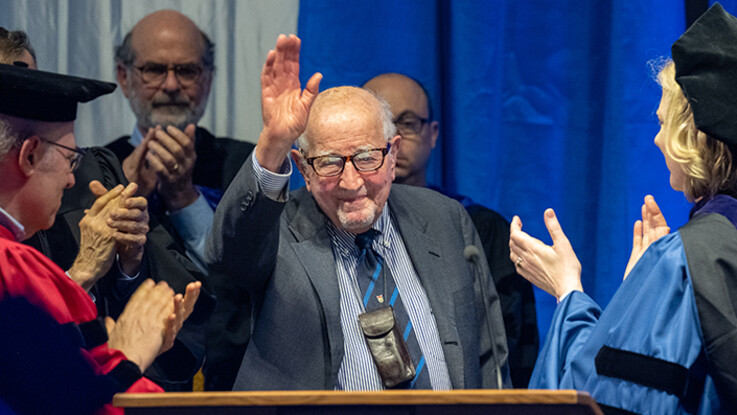 Guido Calabresi waves to the audience from the commencement stage with faculty in academic gowns applauding him