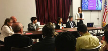 Students sit at a conference table with one female student standing speaking to the audience