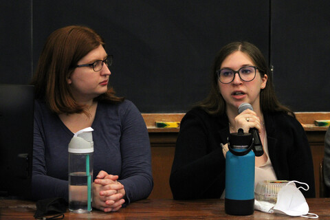 Two students, one with a microphone, seated before a chalk board
