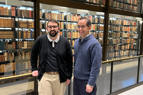 Nicholas Mignanelli and Michael VanderHeijden stand in front a glass walk encasing shelves of lighted books.