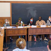 A row of people seated behind a classroom desk, one with a microphone, as students listen