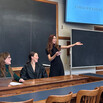 Emilia Wickstead, wearing a black dress, stands in front of a chalkboard and gestures towards the audience. Two women are seated at the table to her left.