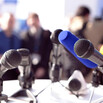 A group of microphones on a table with a crowd in the background.