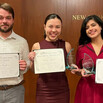 Three students hold up certificates and glass trophies, standing in front of a wood-paneled wall with the words "New York University School of Law"