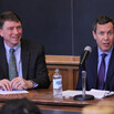 Two men sitting at a speakers’ desk with a microphone in front of a chalkboard