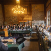 an ornate room with chandelier with many people in suits for a hearing