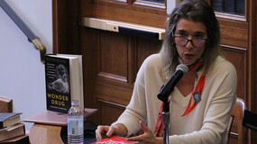 A woman, seated and holding a book, speaks into a microphone.