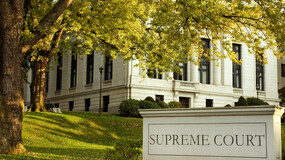 A stone marker with the engraved words “SUPREME COURT” sits in front of a white stone courthouse, surrounded by a lawn and trees 